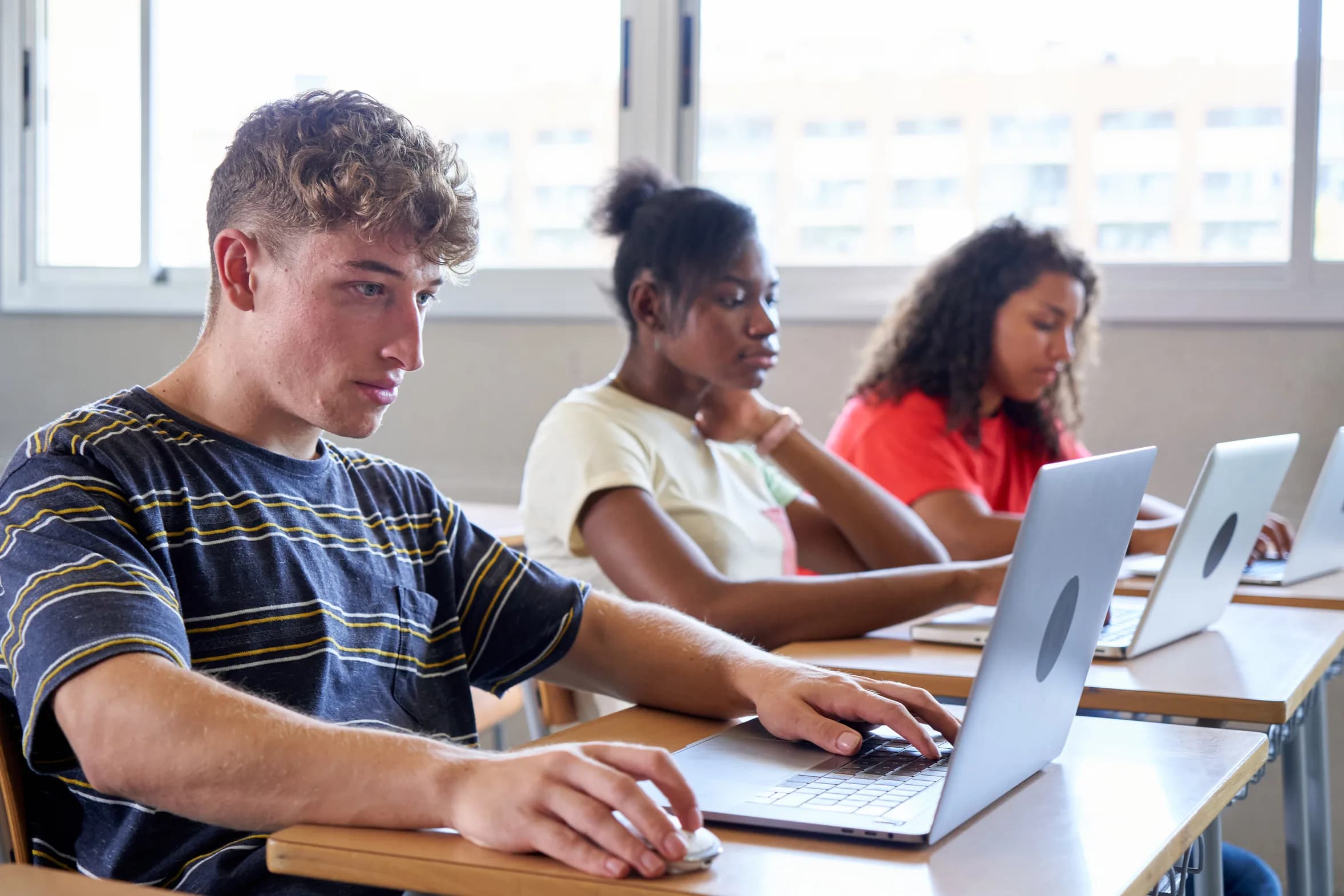 Three teenage students working on laptops at desks in a classroom