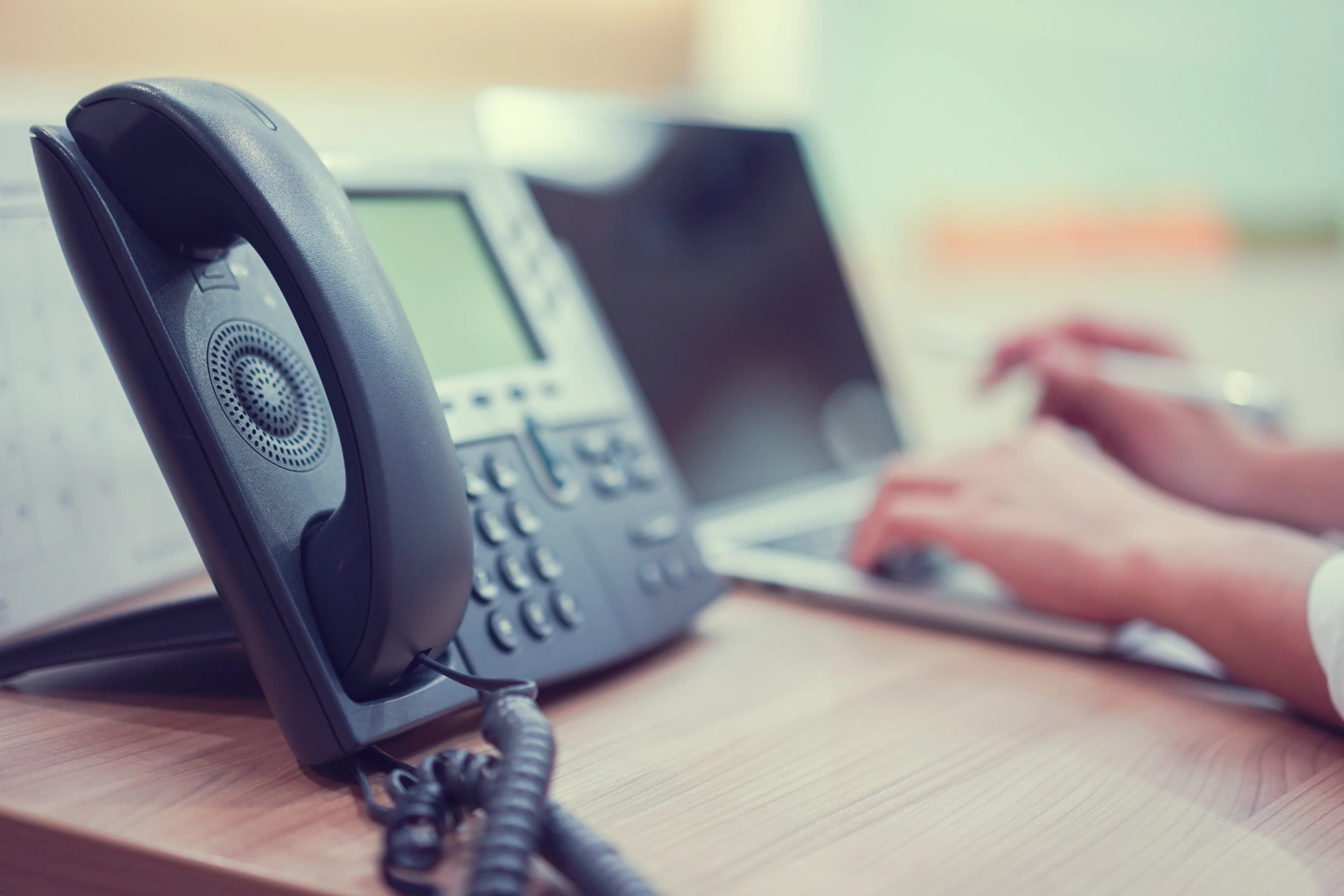 Close-up of a desk phone and an employee working on a laptop at an office desk